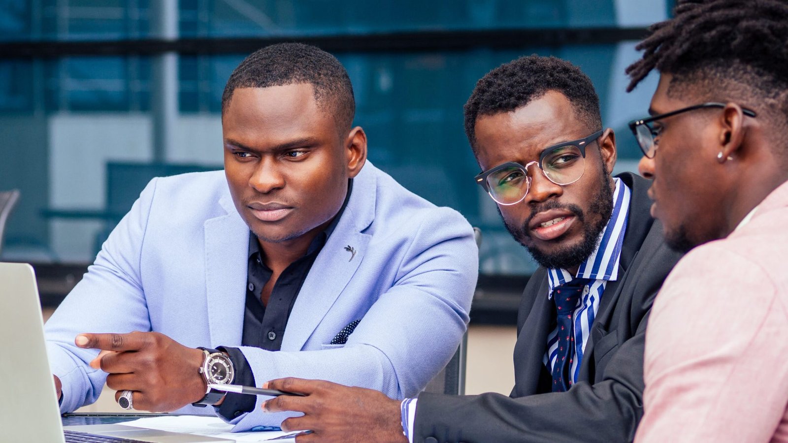 A group of three stylish African American men entrepreneurs in fashion business suits working sitting at table with laptop in a summer cafe outdoors.